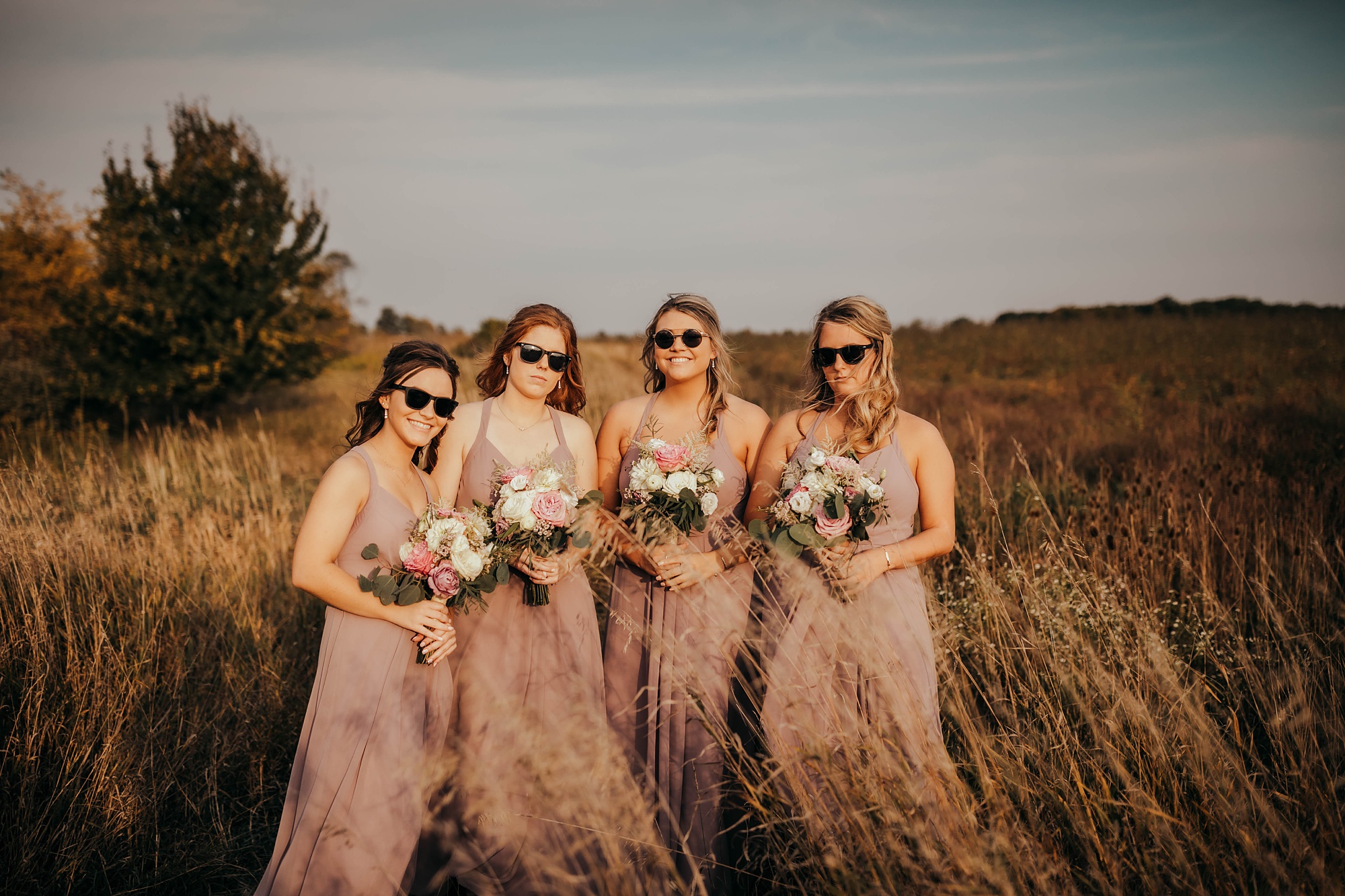 bridesmaids in pink gowns with bouquets pose in fields with sunglasses